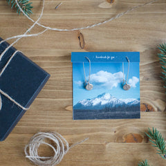 Dainty hand-stamped earrings featuring the state of Alaska. Displayed on a photograph  of the Alaskan mountains